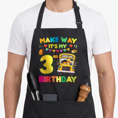 Way birthday apron - Cartoon yellow school bus with children, number 3, and colorful school supplies on canvas fabric — shown on a black cotton canvas apron against a white background