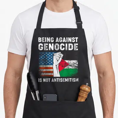 Against palestine apron - Raised fist over US and Palestine flags with text Being Against Genocide Is Not Antisemitism — shown on a black cotton canvas apron against a white background