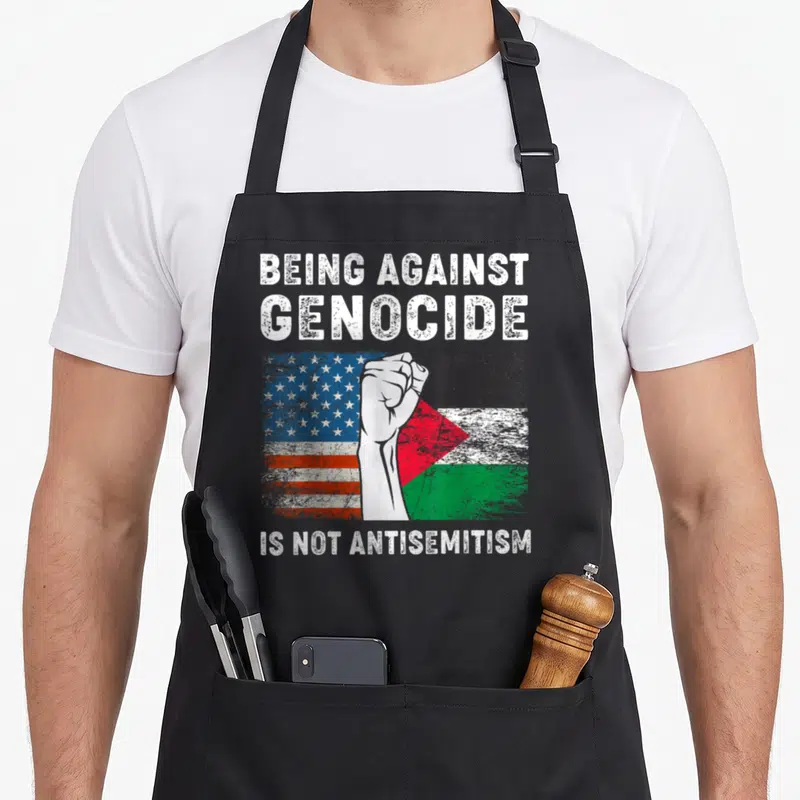 Against palestine apron - Raised fist over US and Palestine flags with text Being Against Genocide Is Not Antisemitism — shown on a black cotton canvas apron against a white background