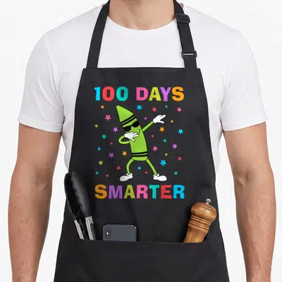 Smarter crayons apron - Dabbing green crayon with sunglasses surrounded by colorful stars and 100 DAYS SMARTER text — shown on a black cotton canvas apron against a white background