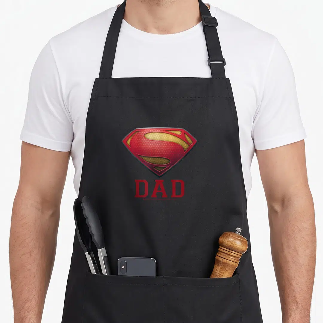 Steel robin dad apron - Realistic red yellow Superman shield above red DAD text on grey background — shown on a black cotton canvas apron against a white background