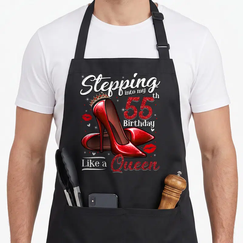 Stepping 55th birthday apron - Red glitter high heels with gold crown and Stepping into my 55th Birthday Like a Queen — shown on a black cotton canvas apron against a white background