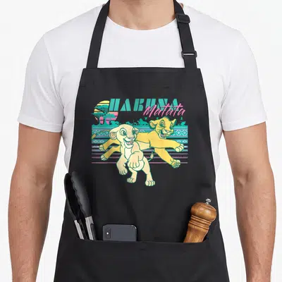 90s lion apron - Two yellow lion cubs running before teal geometric stripes and HAKUNA Matata text — shown on a black cotton canvas apron against a white background