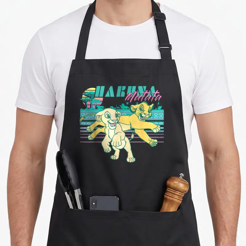 90s lion apron - Two yellow lion cubs running before teal geometric stripes and HAKUNA Matata text — shown on a black cotton canvas apron against a white background