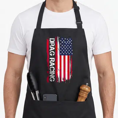 Funny drag racing apron - Vertical DRAG RACING text over a distressed red white and blue USA flag graphic — shown on a black cotton canvas apron against a white background
