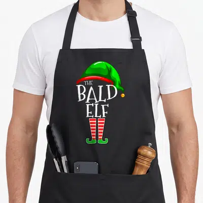 Bald family christmas elf apron - Cartoon elf with green hat, white text, and red striped shoes — shown on a black cotton canvas apron against a white background