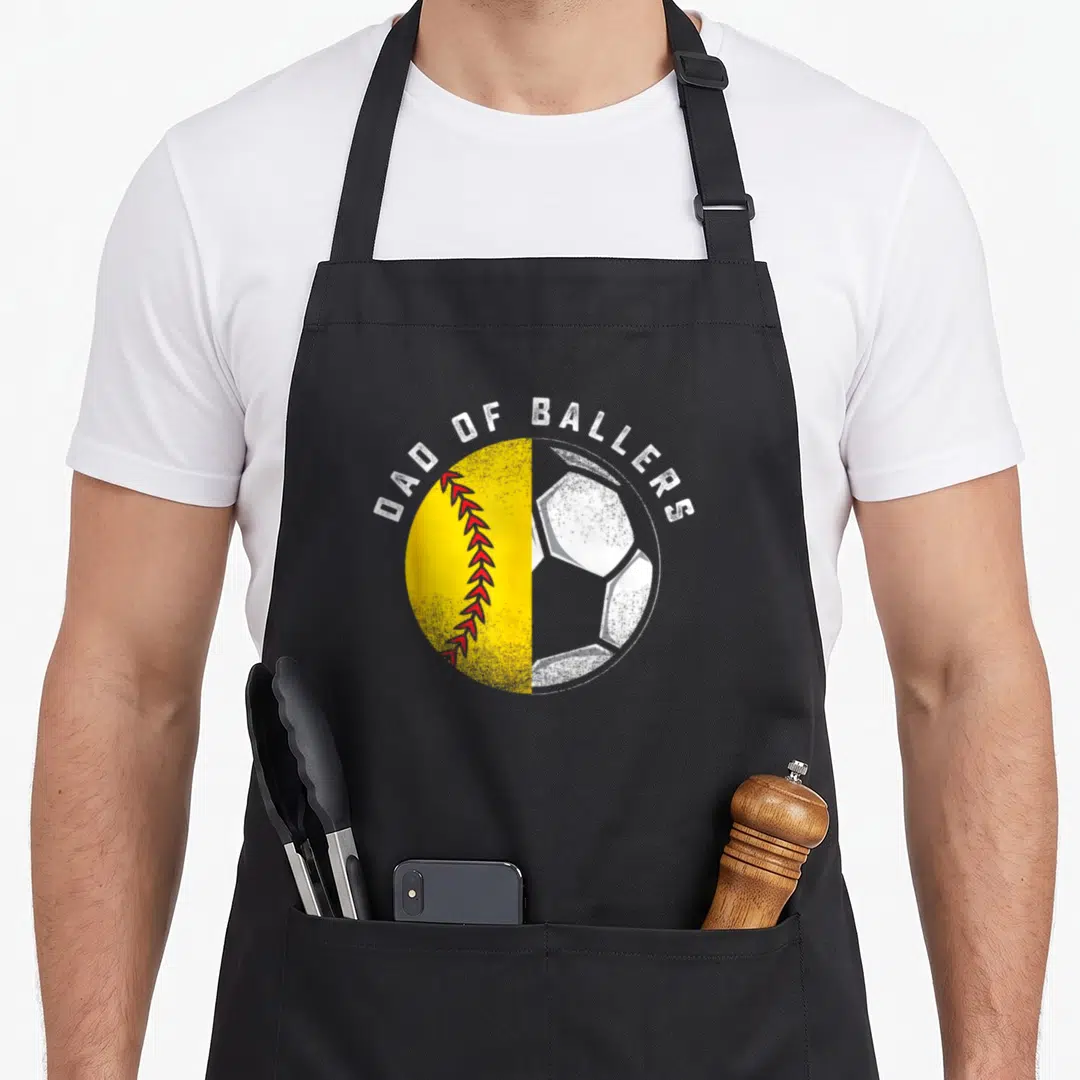 Softball father son apron - Arched Dad of Ballers text over split yellow softball and white soccer ball graphic — shown on a black cotton canvas apron against a white background