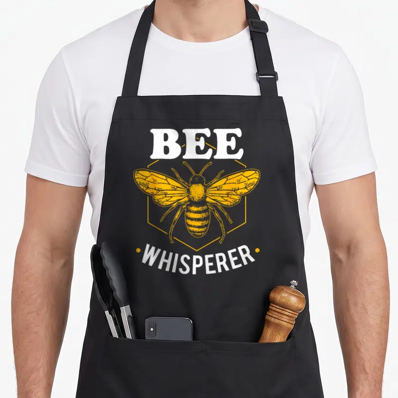 Whisperer bee apron - Yellow bee in hexagon with BEE WHISPERER text in white — shown on a black cotton canvas apron against a white background
