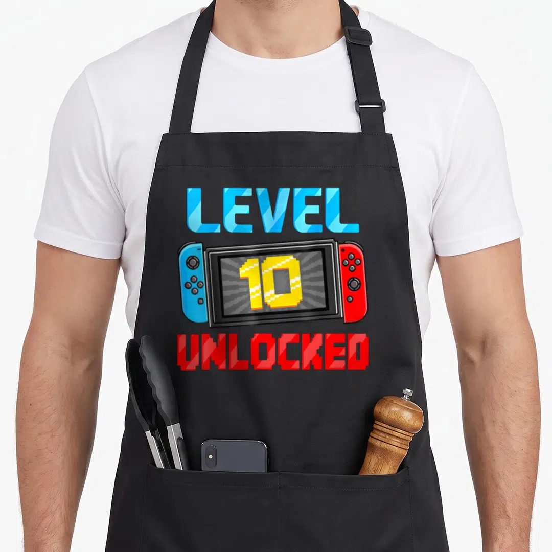 Level unlocked gamer video game apron - Handheld console with blue and red text reading Level 10 Unlocked in pixel art — shown on a black cotton canvas apron against a white background