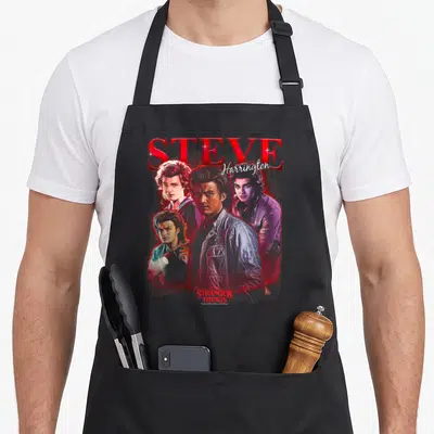 Steve novelty apron - Four portraits of Steve Harrington with red STEVE text and Stranger Things logo — shown on a black cotton canvas apron against a white background