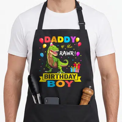 Daddy dinosaur birthday family cooking aprons - Green T-Rex wearing party hat surrounded by balloons and gifts — shown on a black cotton canvas apron against a white background