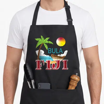 Tourist fiji apron - Cartoon Fiji landscape with sun, palm, boat, and BULA FIJI text in red blue yellow green — shown on a black cotton canvas apron against a white background