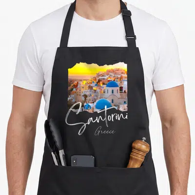 Santorini greece apron - Coastal village photo with blue domes, yellow sunset, and white script reading Santorini Greece — shown on a black cotton canvas apron against a white background