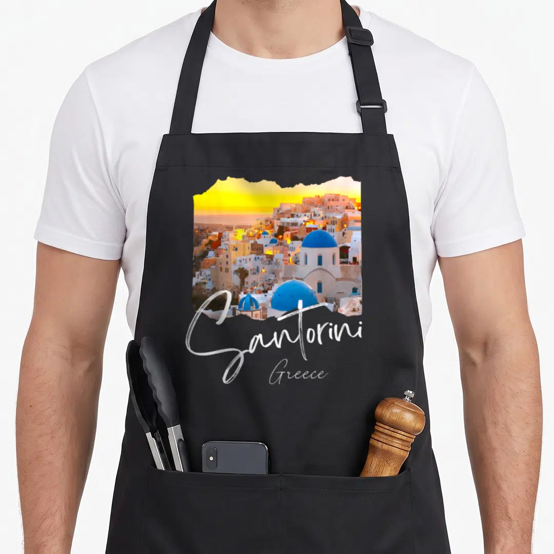 Santorini greece apron - Coastal village photo with blue domes, yellow sunset, and white script reading Santorini Greece — shown on a black cotton canvas apron against a white background