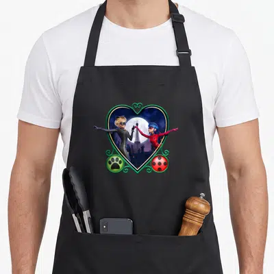 Cat ladybug apron - Two superheroes in a green heart frame with moon, Eiffel Tower, paw print, and ladybug spots — shown on a black cotton canvas apron against a white background