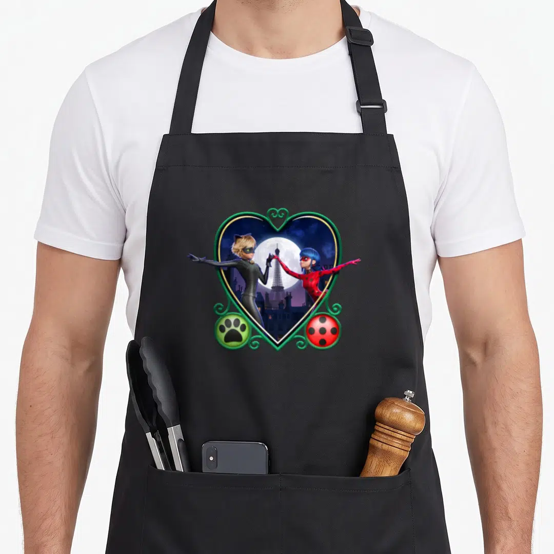 Cat ladybug apron - Two superheroes in a green heart frame with moon, Eiffel Tower, paw print, and ladybug spots — shown on a black cotton canvas apron against a white background