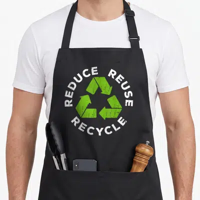 Reduce recycling awareness apron - Distressed green recycling symbol surrounded by white REDUCE REUSE RECYCLE text — shown on a black cotton canvas apron against a white background