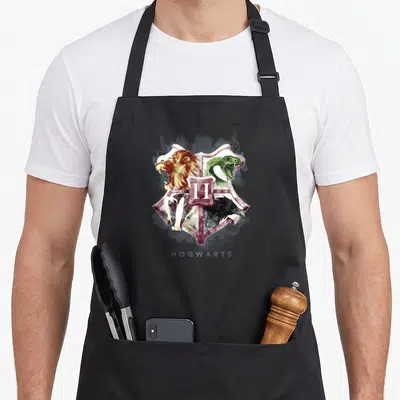 Crest potter apron - Watercolor crest with lion snake badger raven H and HOGWARTS text — shown on a black cotton canvas apron against a white background