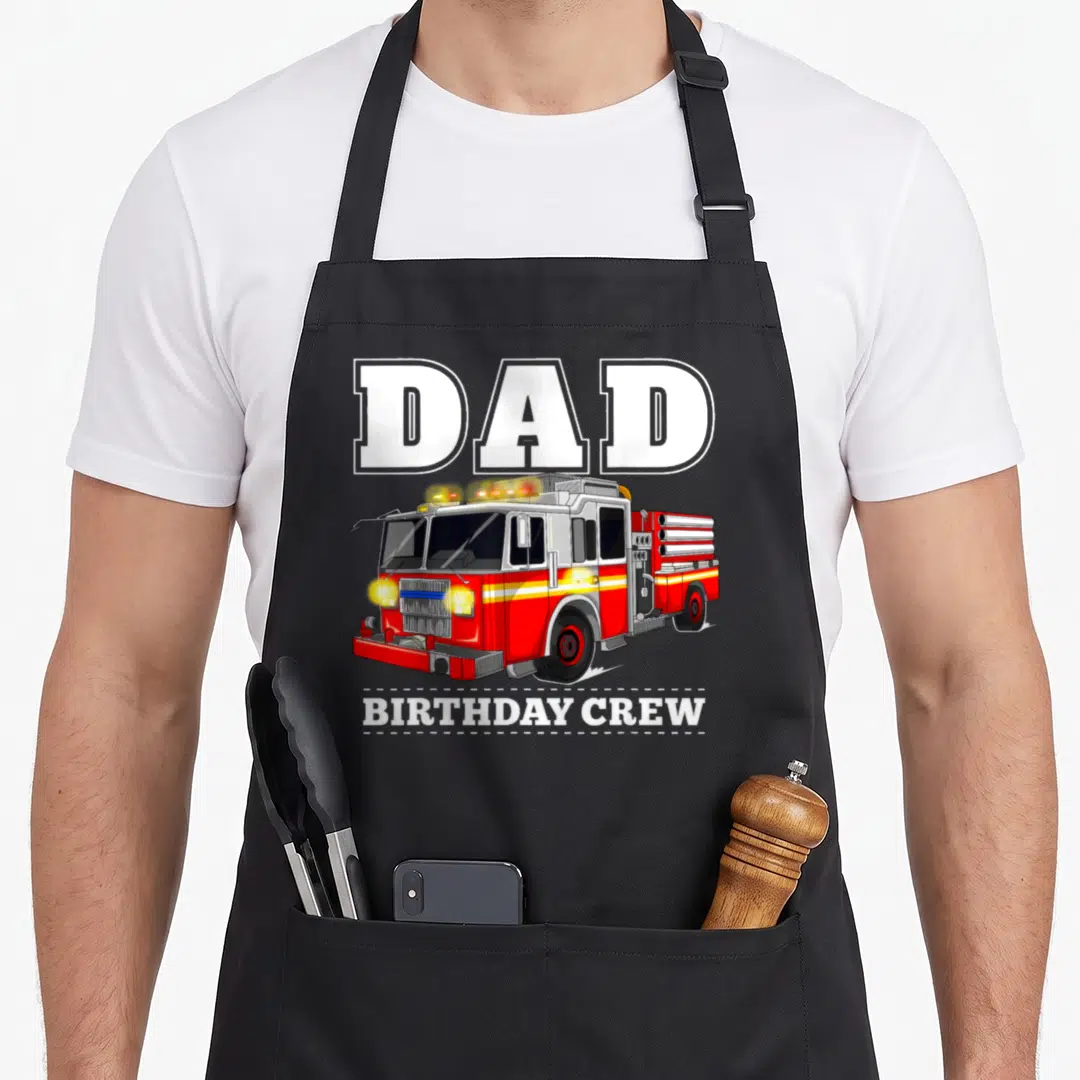 Birthday firefighter aprons - Cartoon red fire truck with Dad Birthday Crew text in white and yellow stripes — shown on a black cotton canvas apron against a white background