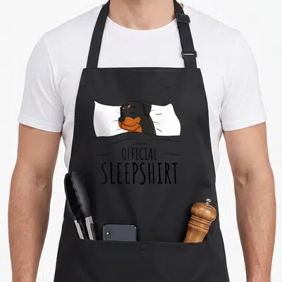 Rottweiler rottie dog apron - Cartoon Rottweiler sleeping on white pillow under grey blanket with official sleepshirt — shown on a black cotton canvas apron against a white background