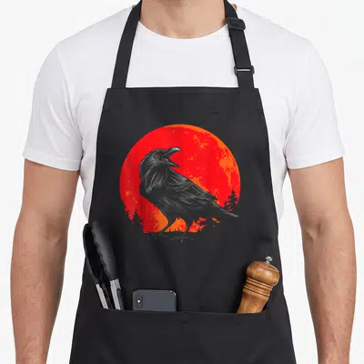 Animal gothic apron - Grey raven on grass in front of red and orange sun with tree silhouettes — shown on a black cotton canvas apron against a white background