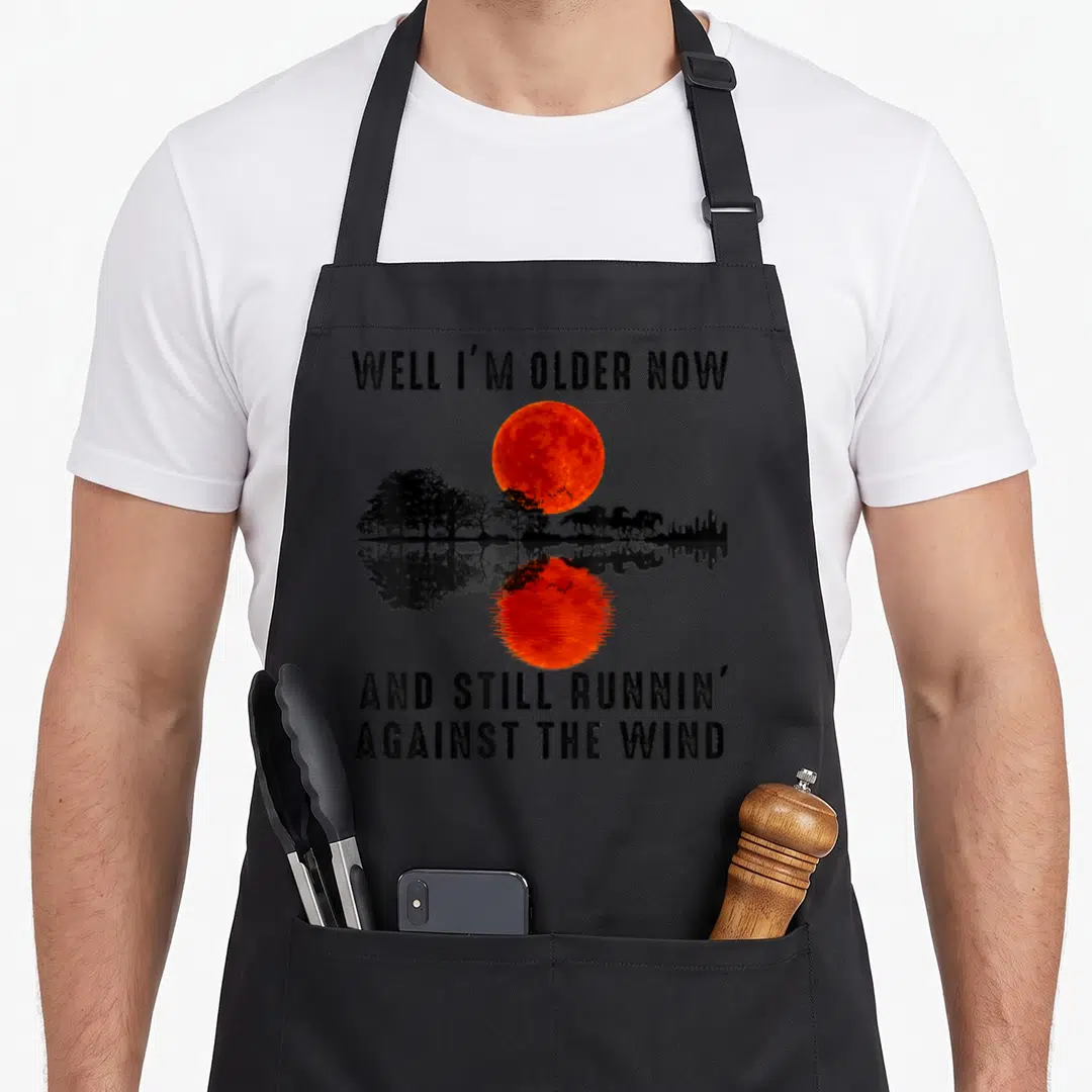 Well running apron - Red moon and black horse silhouettes with text Well I'm Older Now And Still Runnin Against The Wind — shown on a black cotton canvas apron against a white background