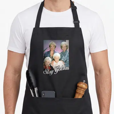 Stay golden apron for dad - Three women in a group photo with Stay Golden script text overlay at the bottom — shown on a black cotton canvas apron against a white background