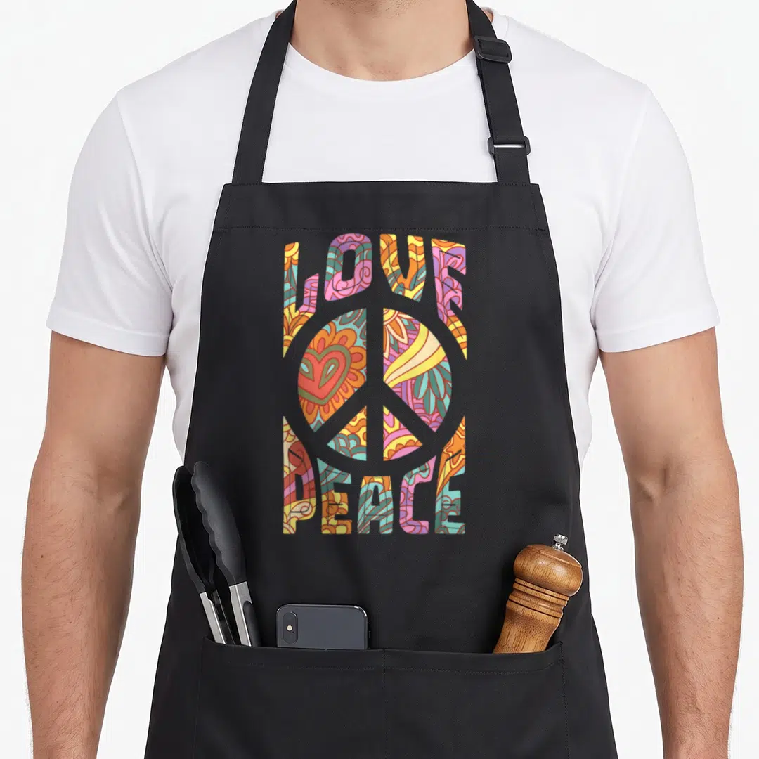 Hippie 60s apron - Retro LOVE and PEACE text surrounding a peace symbol with a heart and floral patterns in orange — shown on a black cotton canvas apron against a white background