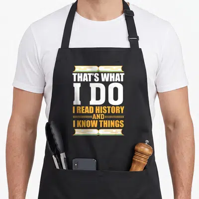Buff teacher apron - Stacked white and orange text reading I read history flanked by open book illustrations — shown on a black cotton canvas apron against a white background