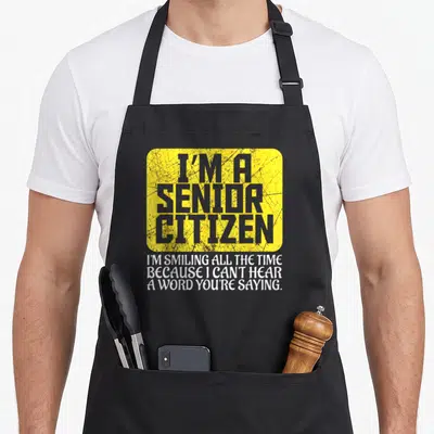 Senior hard of hearing apron - Yellow rectangle with grey text reading I'm a senior citizen above white text about not — shown on a black cotton canvas apron against a white background