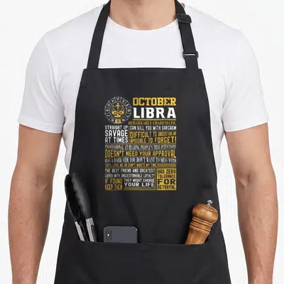 October zodiac aprons - Libra zodiac emblem with scales and stacked yellow and white text listing sarcastic personality — shown on a black cotton canvas apron against a white background