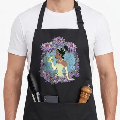 Princess floral apron - Cartoon princess holding frog inside purple lotus flower frame — shown on a black cotton canvas apron against a white background