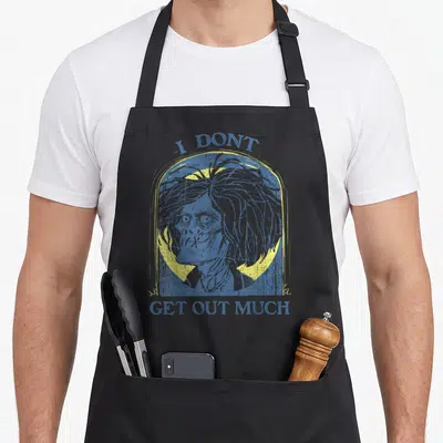 Much billy apron - Distressed blue and yellow skeleton head inside a tombstone outline with text I DONT GET OUT MUCH — shown on a black cotton canvas apron against a white background