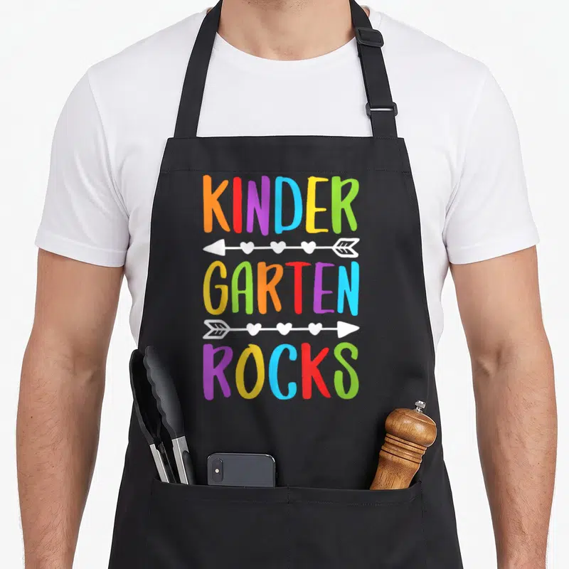 Rock school kindergarten teacher apron - KINDER GARTEN ROCKS text separated by white arrows with hearts in rainbow — shown on a black cotton canvas apron against a white background