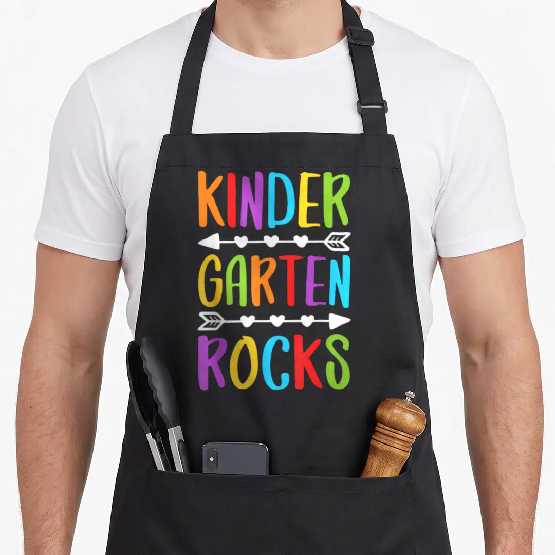 Rock school kindergarten teacher apron - KINDER GARTEN ROCKS text separated by white arrows with hearts in rainbow — shown on a black cotton canvas apron against a white background