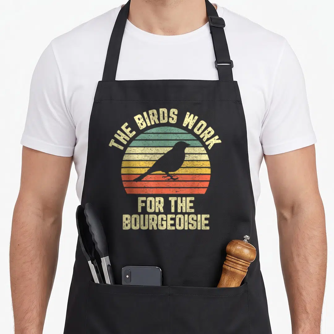 Funny bourgeoisie bird apron - Retro striped sunset with bird silhouette and text THE BIRDS WORK FOR THE BOURGEOISIE — shown on a black cotton canvas apron against a white background