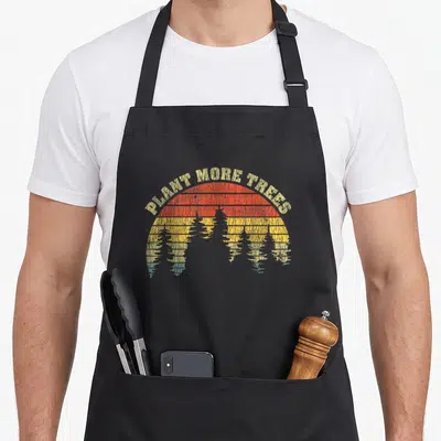 Plant change apron - PLANT MORE TREES text arching over striped sun with tree silhouettes in red and yellow — shown on a black cotton canvas apron against a white background