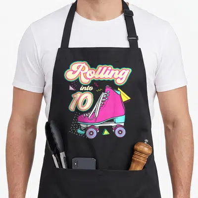 Skate 10th birthday apron - Pink roller skate with Rolling into 10 text and geometric confetti shapes — shown on a black cotton canvas apron against a white background