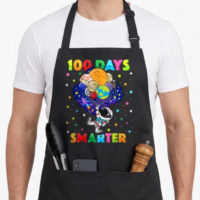 Smarter astronaut space apron - Cartoon astronaut holding planet balloons with 100 DAYS SMARTER rainbow text — shown on a black cotton canvas apron against a white background