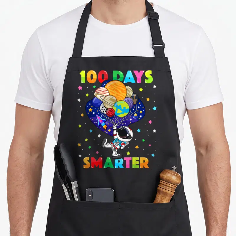 Smarter astronaut space apron - Cartoon astronaut holding planet balloons with 100 DAYS SMARTER rainbow text — shown on a black cotton canvas apron against a white background