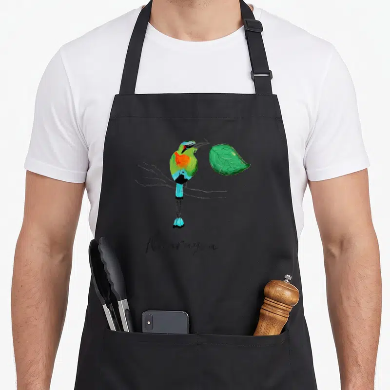 Nicaragua colorful aprons - Watercolor bird perched on branch holding green leaf with Nicaragua text below — shown on a black cotton canvas apron against a white background