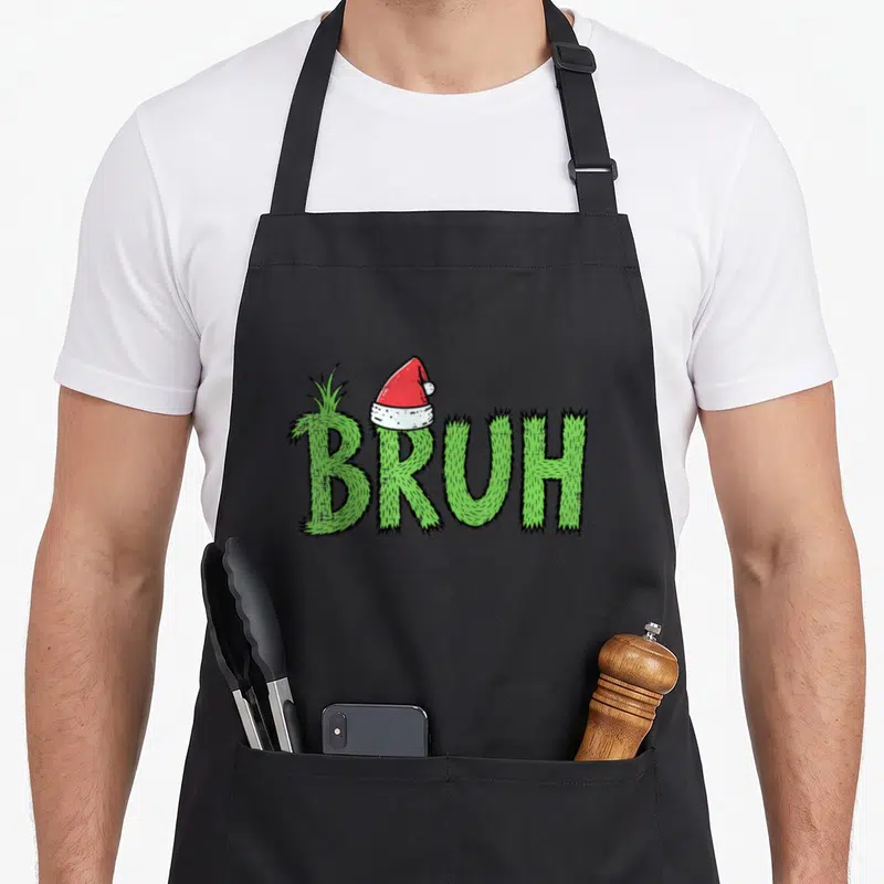 Bruh santa aprons - Green grass textured text reading BRUH with a red Santa hat on the B letter — shown on a black cotton canvas apron against a white background