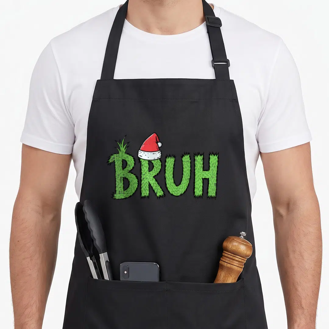 Bruh santa aprons - Green grass textured text reading BRUH with a red Santa hat on the B letter — shown on a black cotton canvas apron against a white background