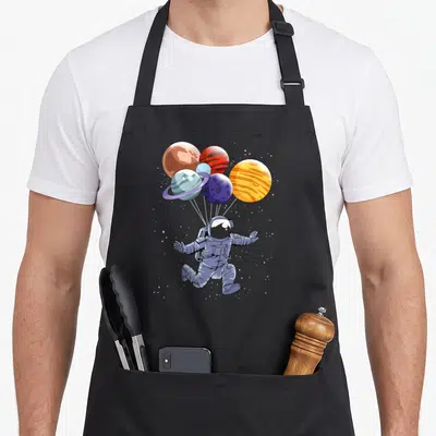 Space balloon apron - Cartoon astronaut floating below six colorful planet balloons connected by white strings amidst — shown on a black cotton canvas apron against a white background