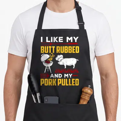 Butt pulled addict apron - Pig in chef hat next to grill with text I like my butt rubbed and my pork pulled — shown on a black cotton canvas apron against a white background
