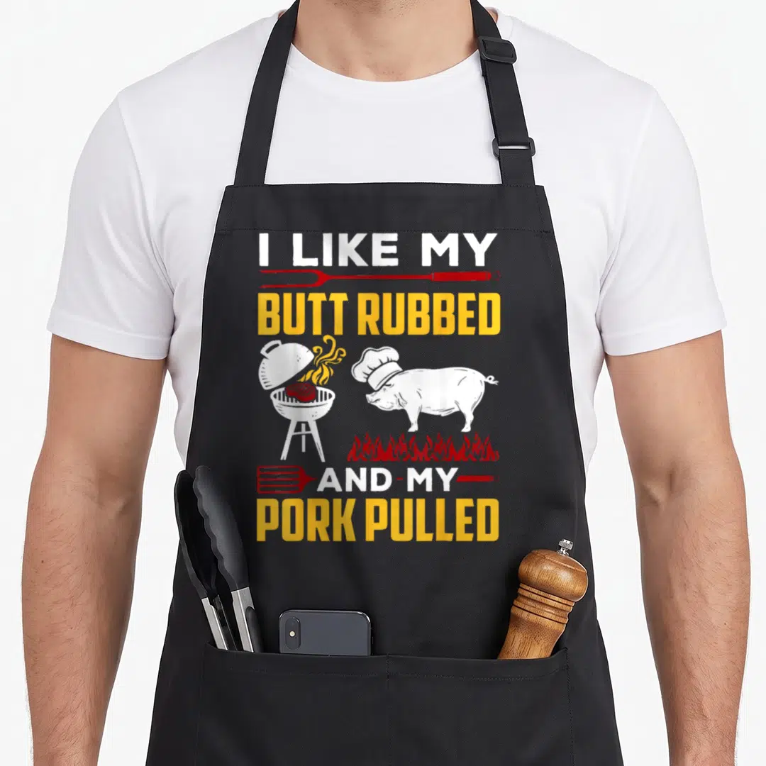 Butt pulled addict apron - Pig in chef hat next to grill with text I like my butt rubbed and my pork pulled — shown on a black cotton canvas apron against a white background