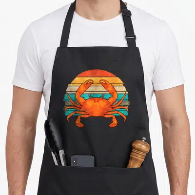 Seafood crabbing crab apron - Retro orange crab centered on a striped semi-circle sun in blue and yellow — shown on a black cotton canvas apron against a white background