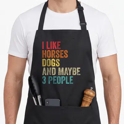 Horse dog apron - Stacked colorful text reading I Like Horses Dogs And Maybe 3 People in red orange yellow beige teal — shown on a black cotton canvas apron against a white background