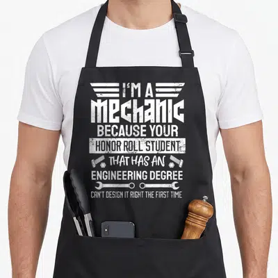Diy garage apron - White distressed text reading I'm a mechanic with two wrenches and screws — shown on a black cotton canvas apron against a white background