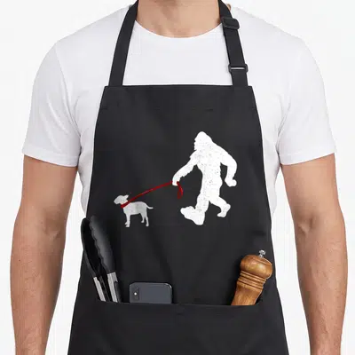 Bigfoot dog dad apron - White Bigfoot silhouette walking a small dog on a red leash with distressed texture — shown on a black cotton canvas apron against a white background